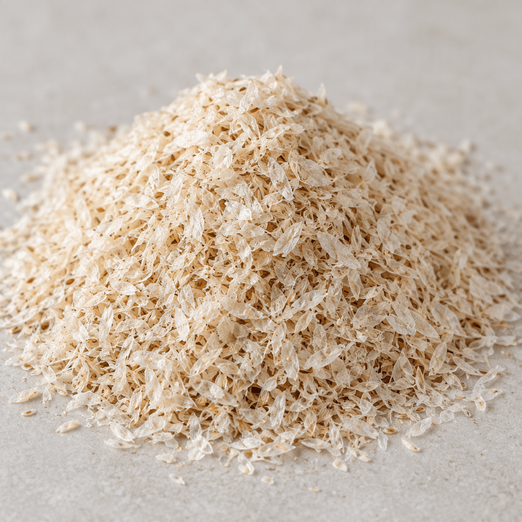 A small mound of dry psyllium husk consists of light beige, fine, papery flakes and tiny curled fragments.