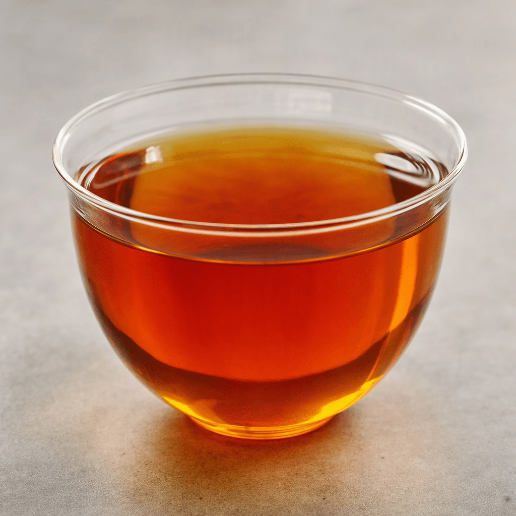 Clear amber-gold tea with a reddish glow sits in a transparent glass cup, its surface smooth and free of visible leaves or additions.