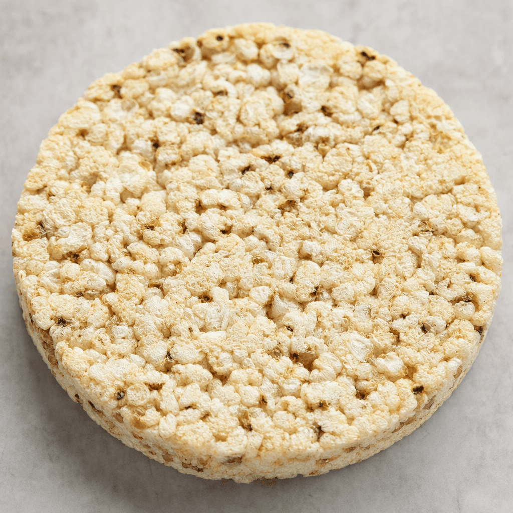A round, flat, lightly textured cake of puffed grains, pale beige with scattered darker buckwheat flecks and an uneven, porous surface.