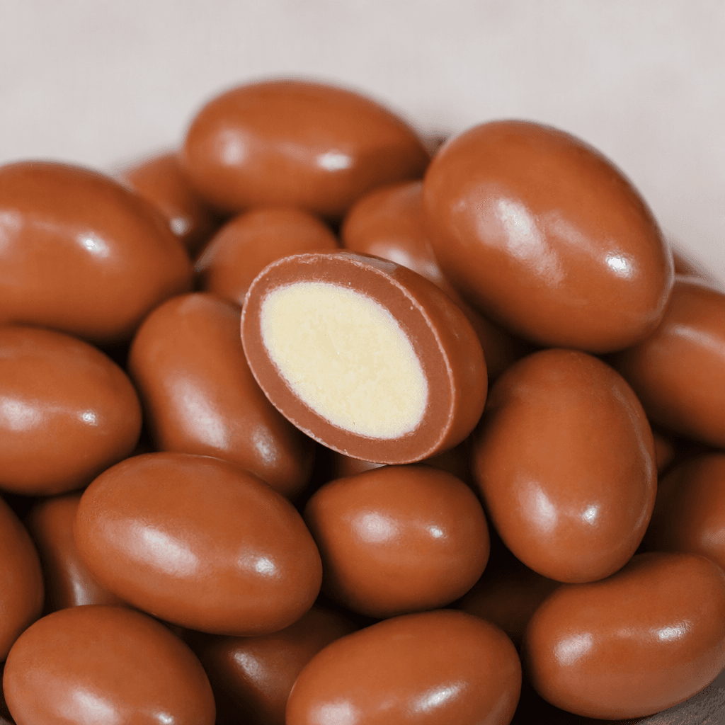 Small, smooth, oval milk-chocolate pieces with a glossy medium-brown coating are arranged together, with one cut open to show its pale, creamy filling.