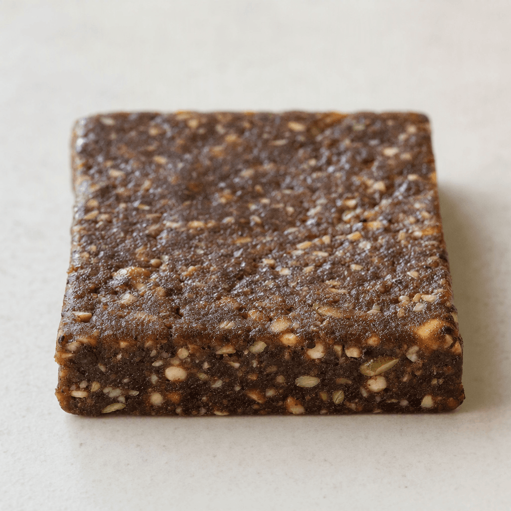 A compact, rectangular dark-brown bar with a dense, slightly uneven surface and small pieces of nuts and seeds visible throughout.