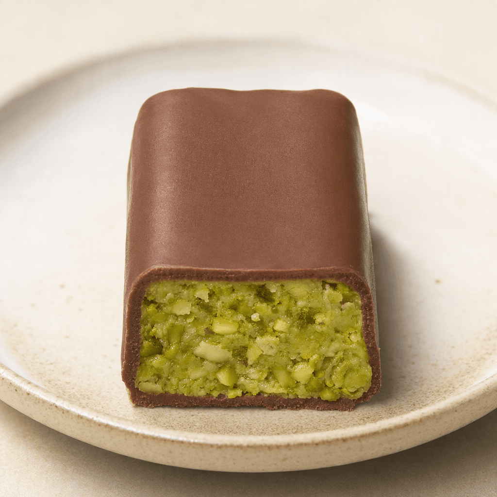 A small rectangular chocolate-coated bar with a smooth brown surface and a pale green pistachio filling flecked with finely chopped nuts visible at the cut end, served on a plain plate.