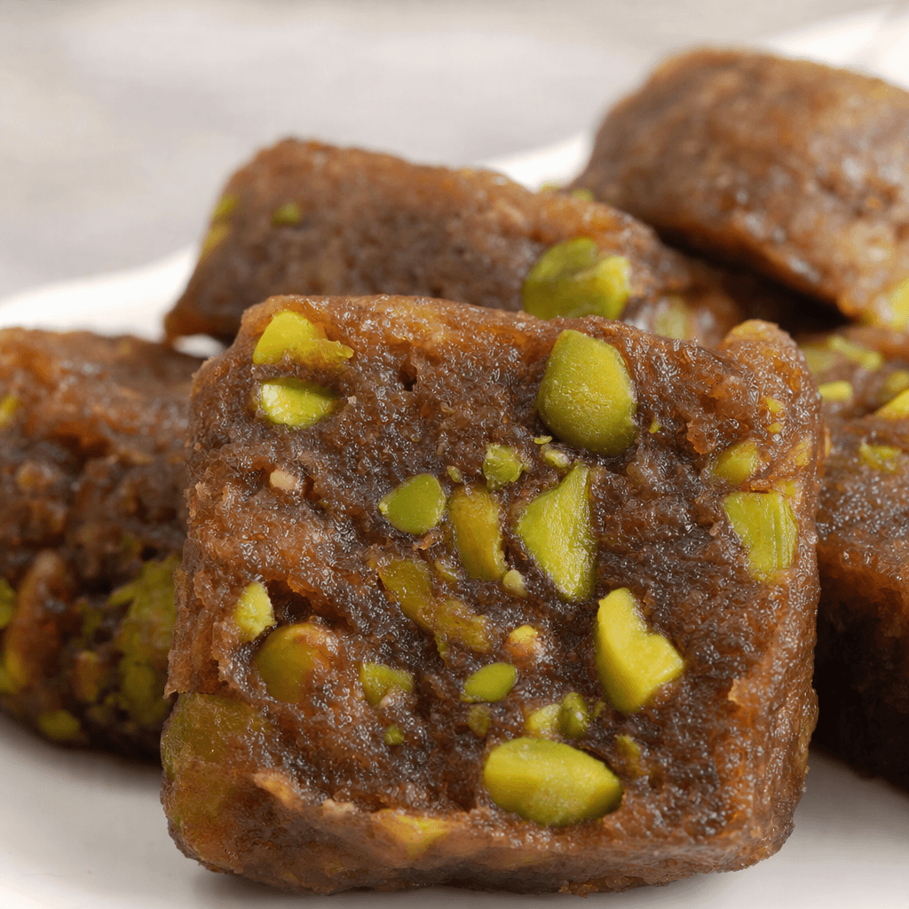 A dense, dark caramel-brown date halva is cut into thick pieces, with bright green pistachio fragments scattered through its slightly coarse, compact interior.