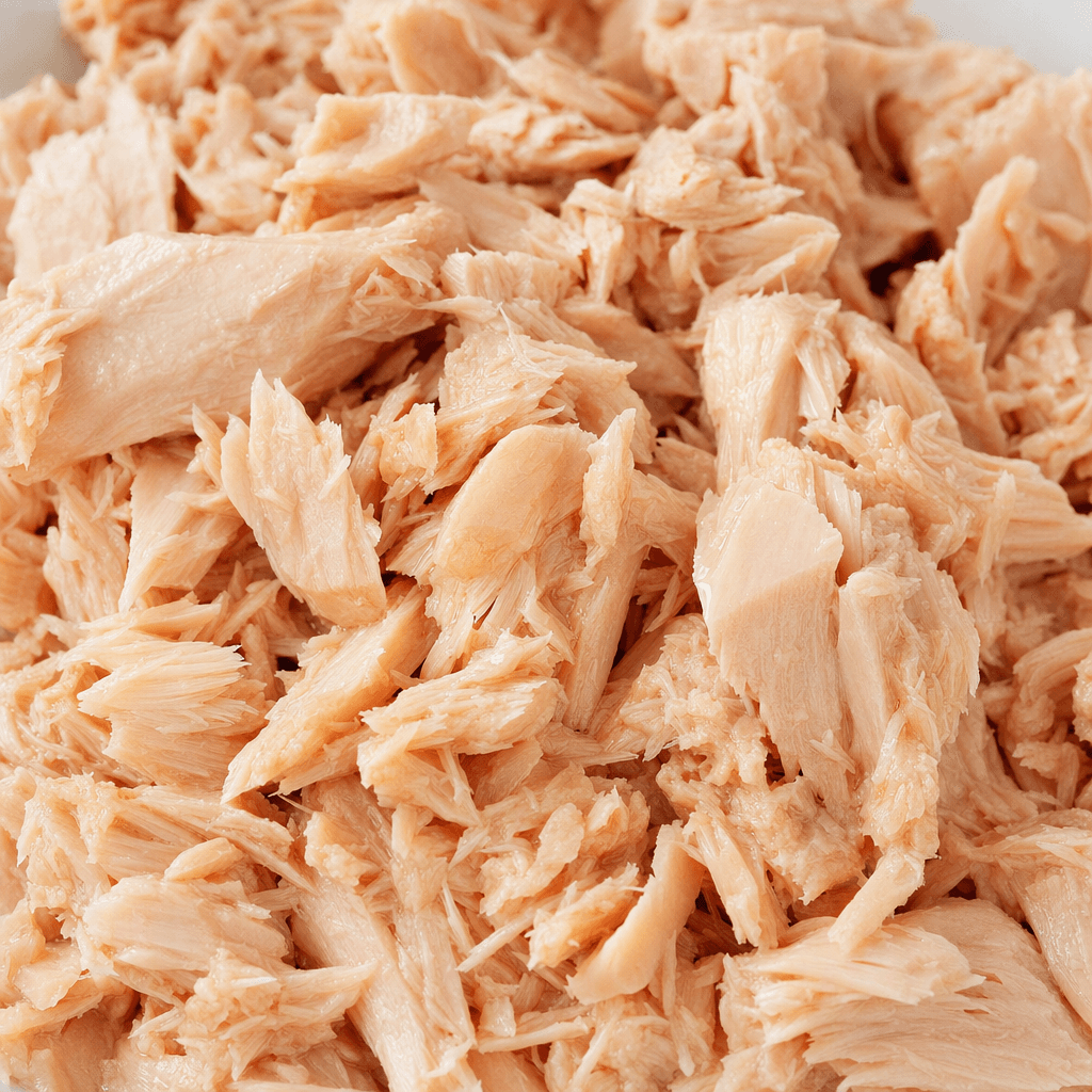 Moist, pale pink-beige tuna flakes and small fibrous chunks are piled together, with a slight sheen of oil.