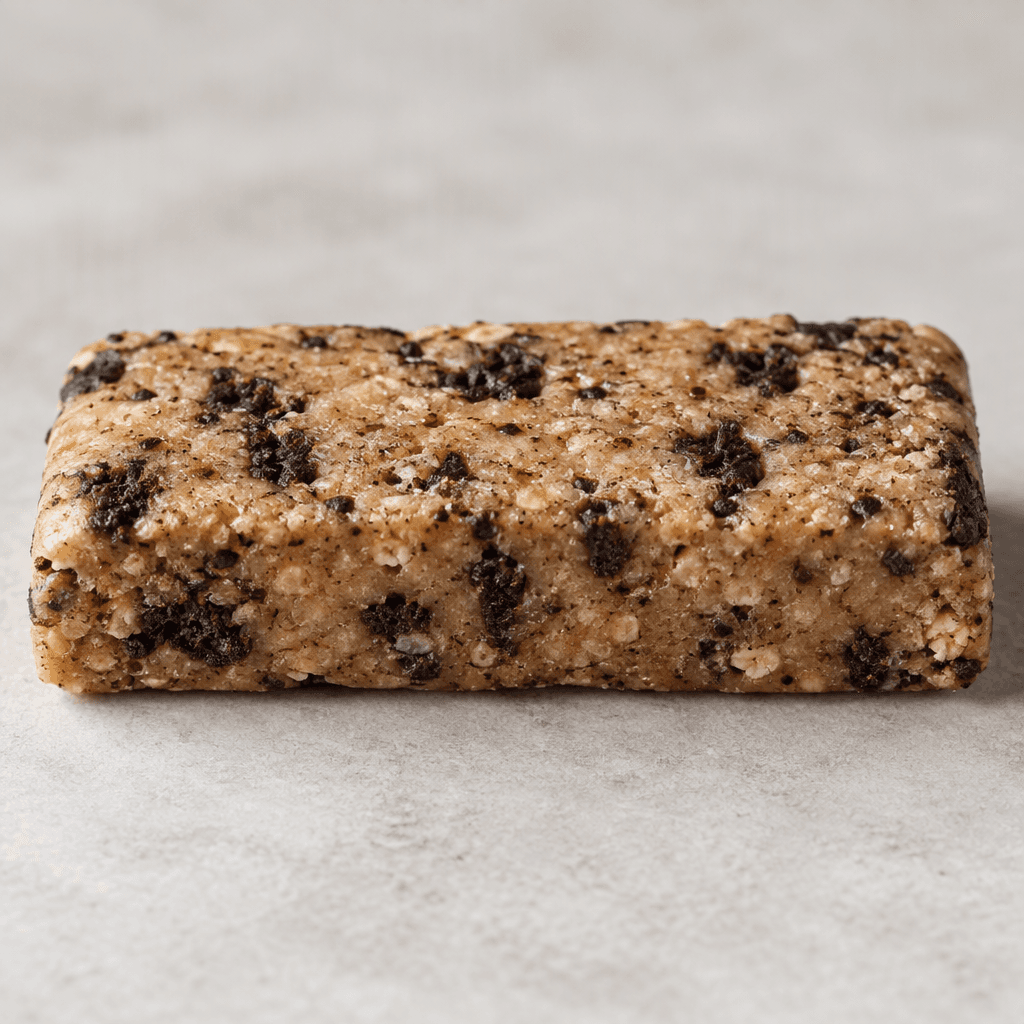 A compact, rectangular tan-brown protein bar with a slightly uneven surface and scattered darker cookie crumbs, shown whole on a plain surface.