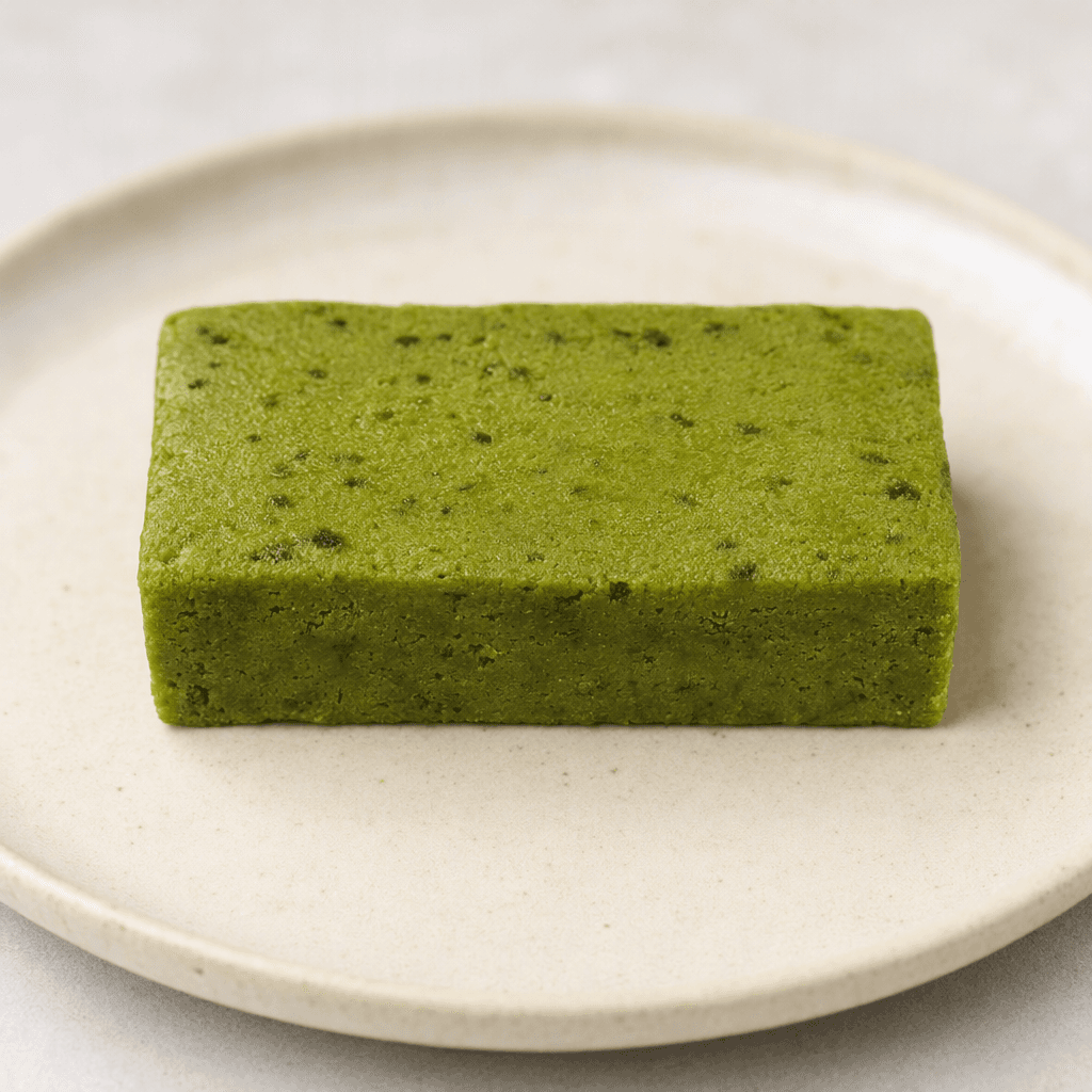 A compact rectangular matcha-green bar with a firm, slightly uneven surface and small darker green flecks, shown whole on a plain plate.