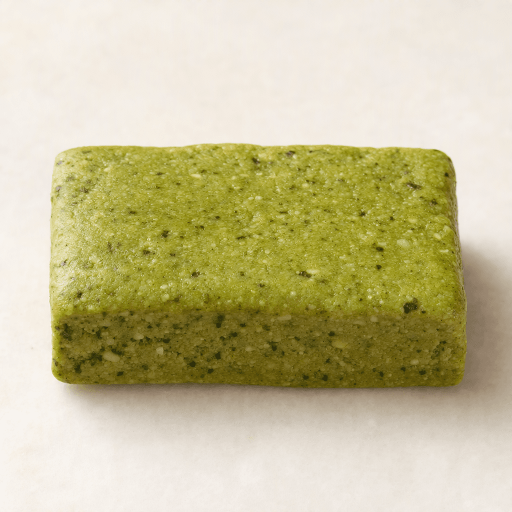 A compact, rectangular matcha protein bar with a muted pale-green surface, fine darker-green flecks, and a slightly uneven, dense-looking texture.