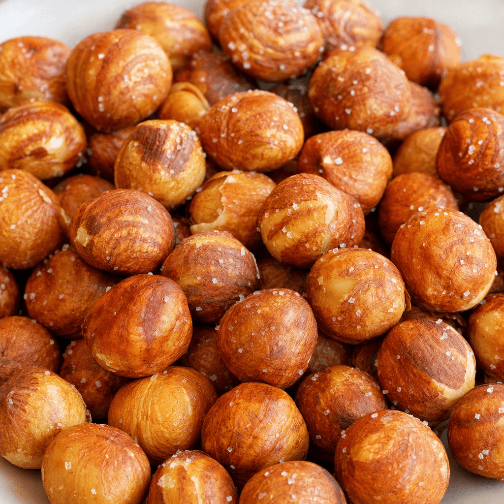 Whole round-to-oval hazelnut kernels with mottled golden-brown skins and a light scattering of visible salt crystals are piled together.