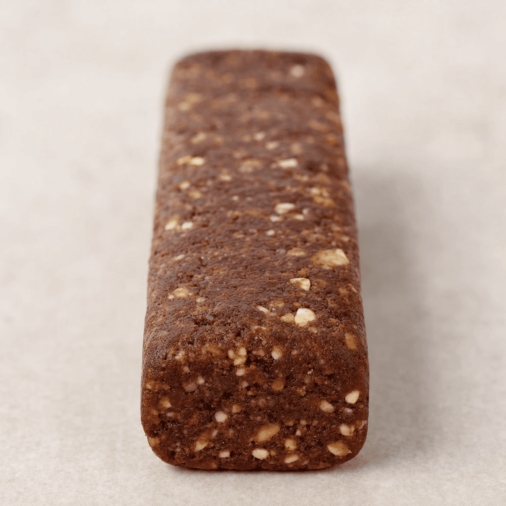 An elongated, rectangular protein bar with rounded edges, a cocoa-brown surface, and small pale and dark pieces visible throughout its dense, slightly rough texture.
