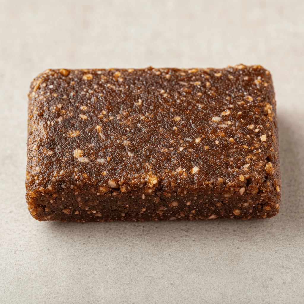 A small, rectangular, dark-brown protein bar with a slightly uneven, compact surface and a few visible lighter flecks.