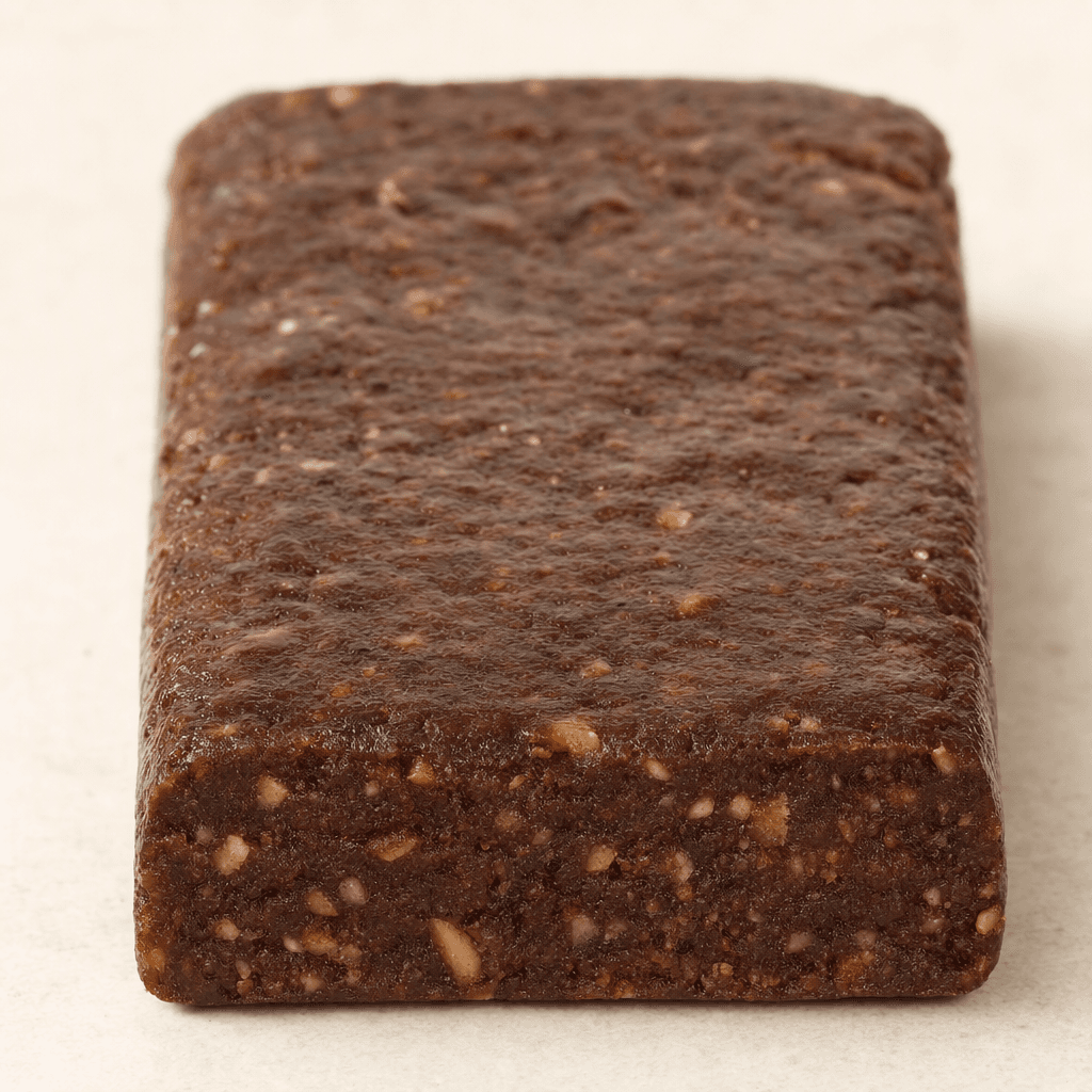 A compact, rectangular protein bar with a dark brown, slightly uneven surface and a dense, chewy-looking interior visible along one edge.