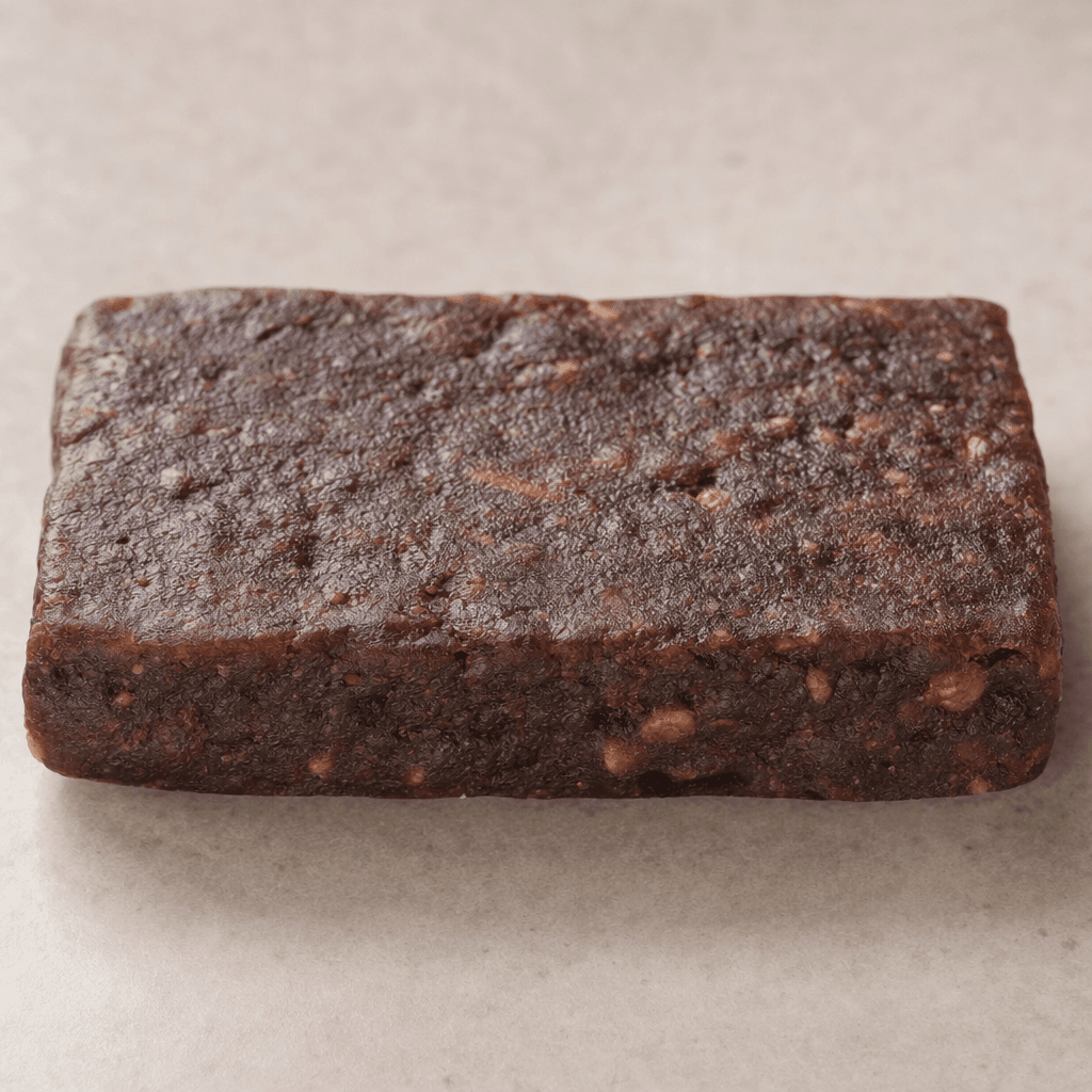 A compact, rectangular chocolate-brown bar with a slightly uneven, dense-looking surface and a few lighter brown bits visible.