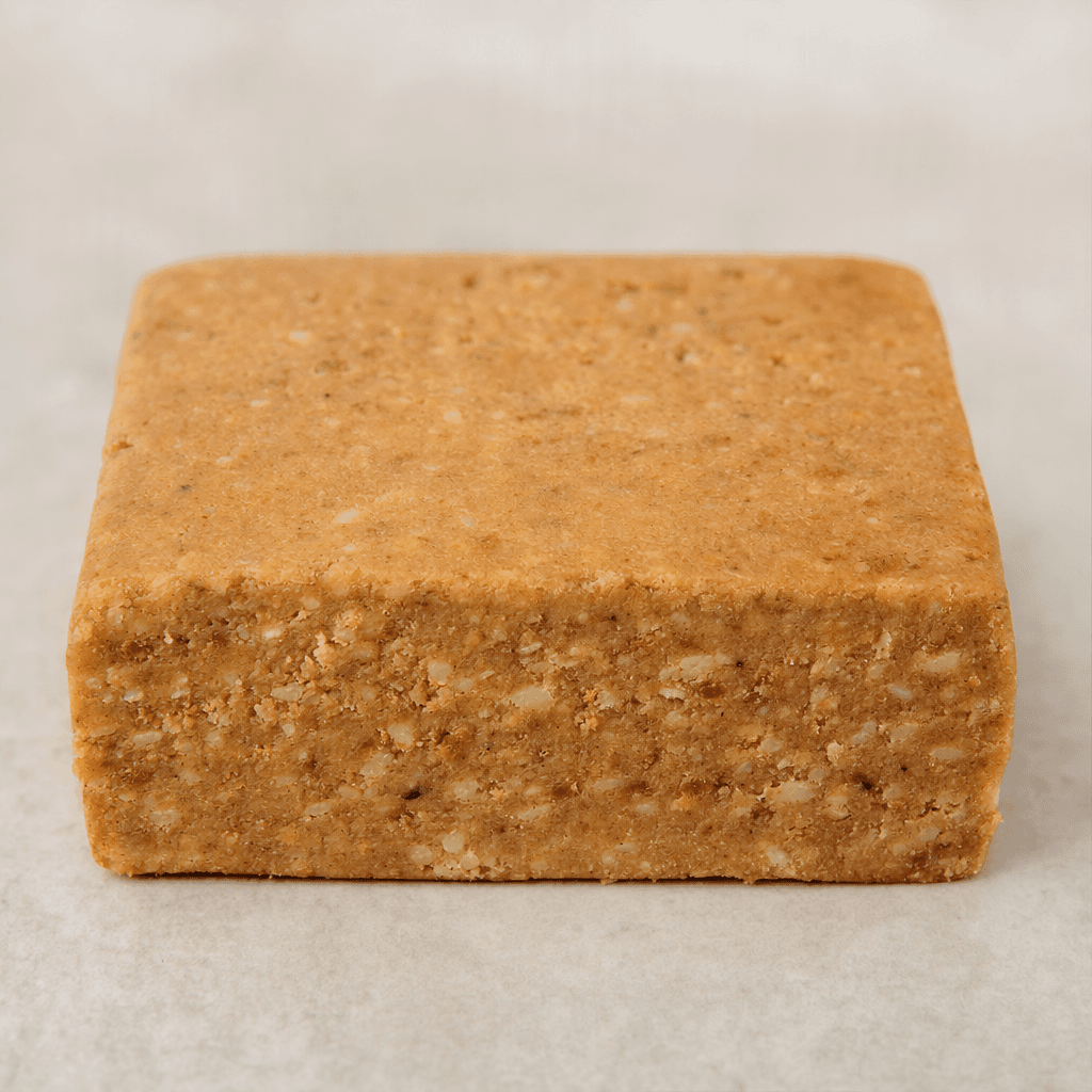 A compact rectangular protein bar with a matte tan-brown surface, slightly uneven edges, and small pale and darker crumb-like flecks visible across its dense cut face.