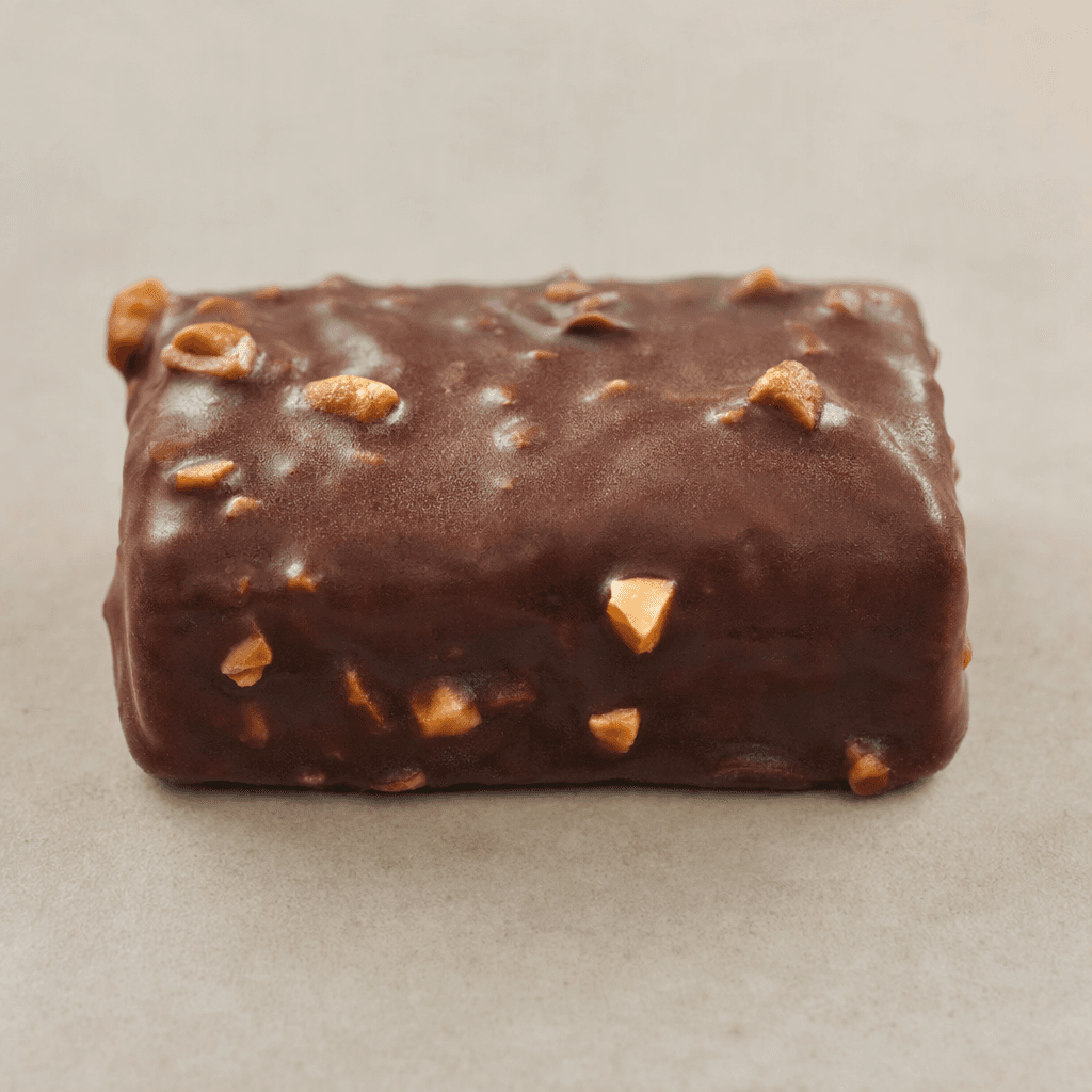 A compact rectangular bar with a dark-brown chocolate coating, a slightly uneven surface, and small hazelnut pieces visible on top and along the edges.