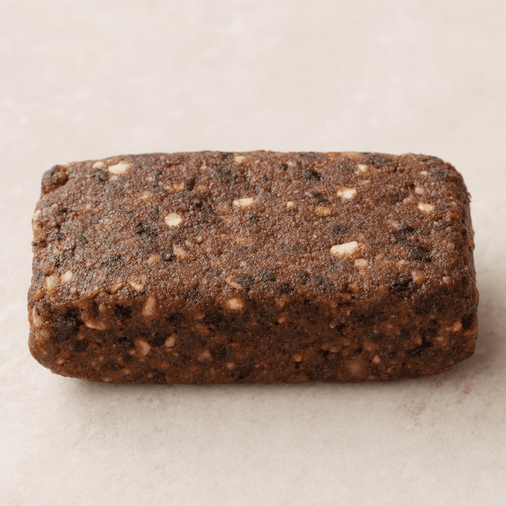 Tek başına sunulan, yüzeyi hafif pütürlü ve içinde küçük parçacıklar görünen, kompakt dikdörtgen biçimli kahverengi bir protein barı.