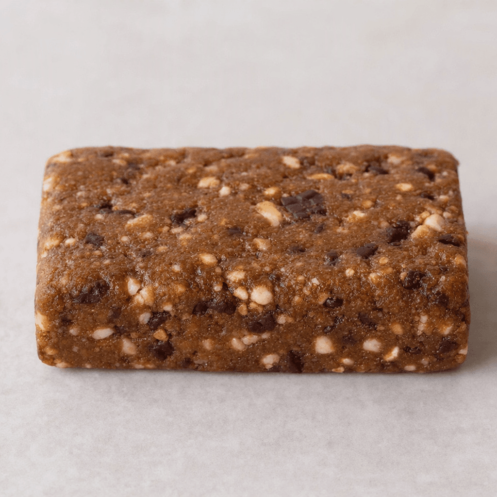 A compact rectangular brown protein bar with a slightly uneven, dense surface and small pale and dark pieces visible throughout, resting on a plain surface.