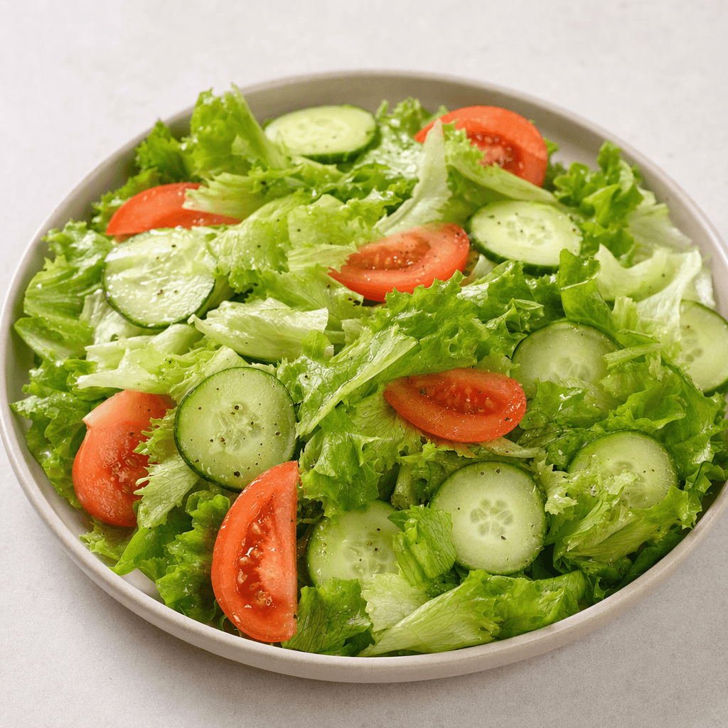 A shallow plate holds crisp green lettuce leaves with thin cucumber slices and red tomato wedges, lightly glistening with a small amount of dressing.