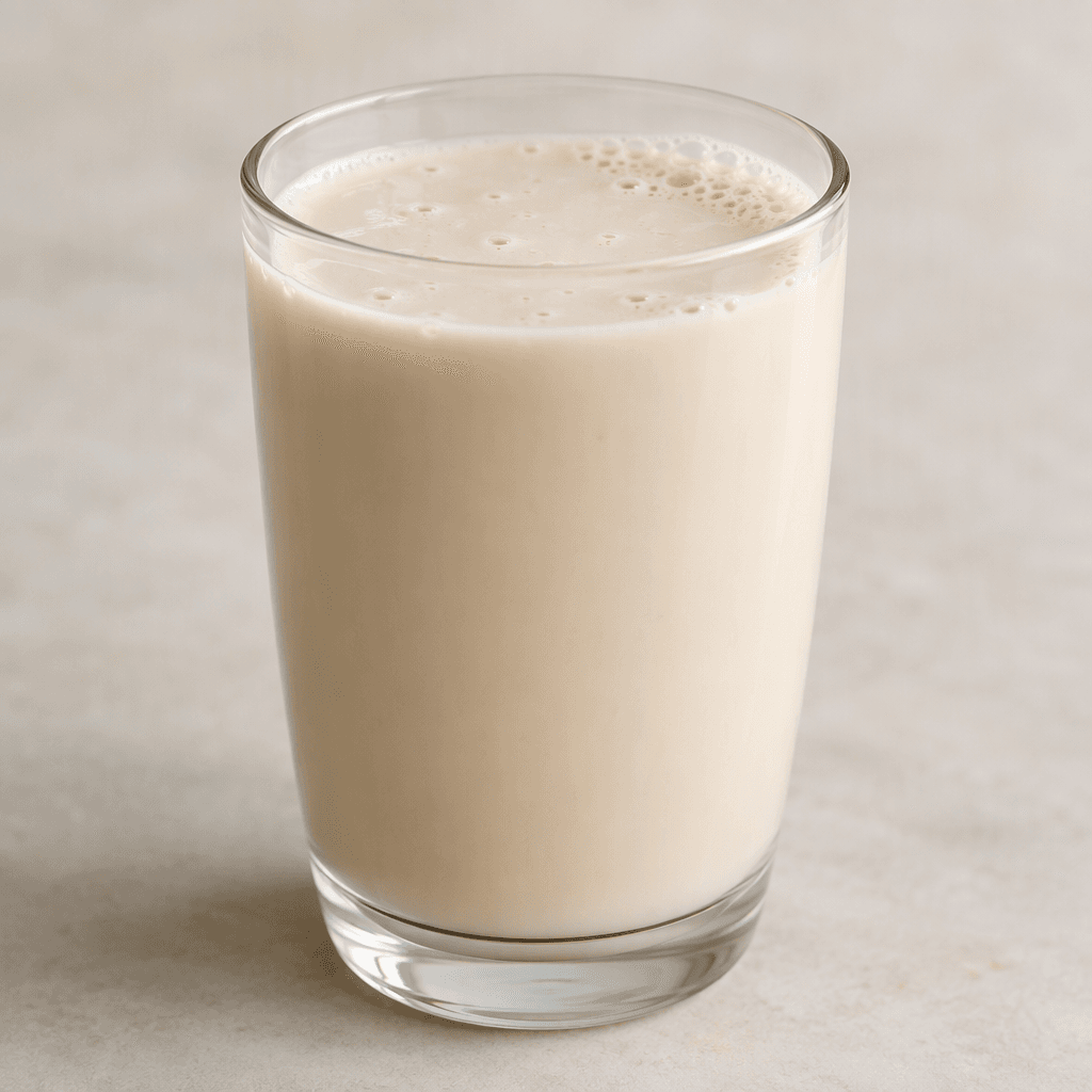 Opaque, creamy beige oat milk fills a clear glass, with a smooth pourable texture and a few tiny bubbles on the surface.
